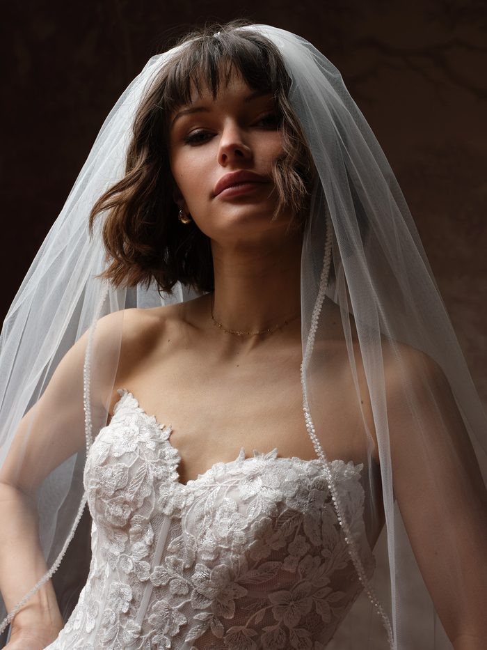 Close-up of the Essentials Edit fingertip veil, featuring soft ivory tulle with a delicate beaded edge draped over a bridal look with a floral lace bodice. Detailed view showcases the veil’s lightweight texture and elegant finishing details.