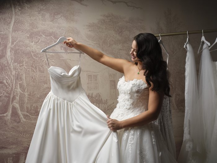 Person wearing the Benton wedding dress tries on another gown from a bridal rack in a boutique-inspired setting. The Benton gown features a strapless bodice with dimensional floral lace appliqués and a full tulle skirt, while the person holds up a satin A-line wedding dress with a draped sweetheart neckline for comparison.