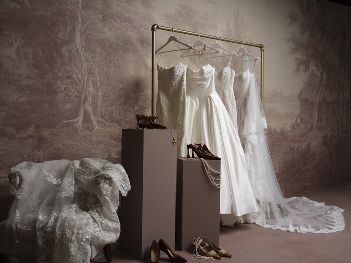 A curated bridal display featuring three wedding gowns hanging on a gold clothing rack, surrounded by bridal accessories including heels, jewelry, and lace fabric. The gowns showcase a variety of silhouettes and design details, including satin and lace fabrics, against a vintage-inspired mural backdrop.