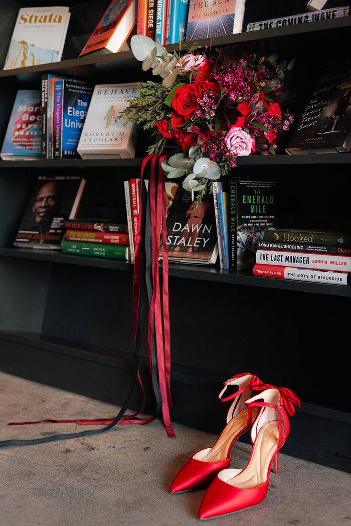 Halloween-inspired bridal details featuring a dramatic bouquet of red, burgundy, and blush blooms with flowing black and burgundy ribbons arranged on a bookshelf, accented by bold red satin heels for a romantic gothic wedding aesthetic.