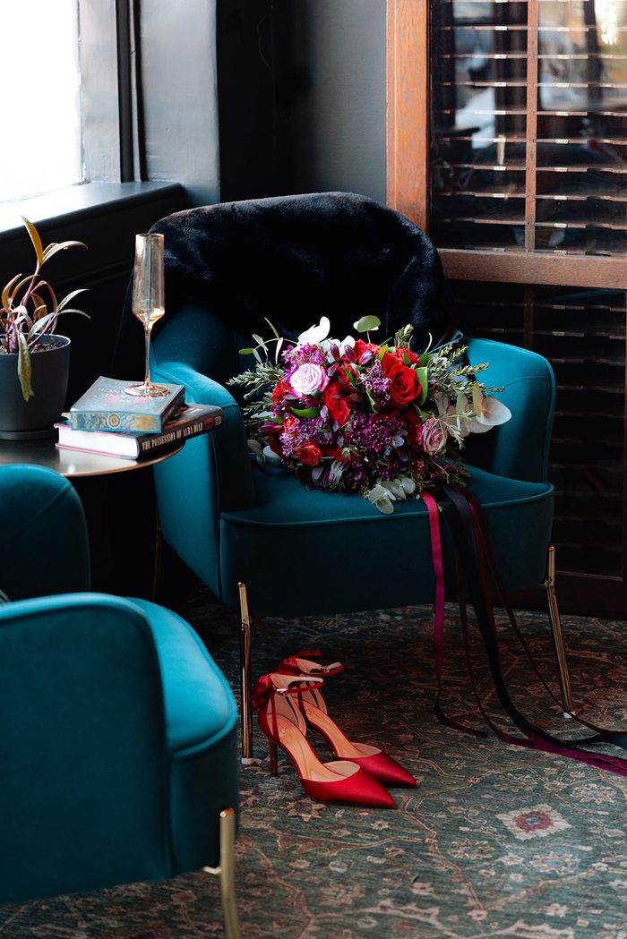 Moody Halloween wedding inspiration featuring a lush bouquet of red, burgundy, and blush flowers with trailing black and burgundy ribbons displayed on a teal velvet chair, paired with vibrant red satin heels in a dark, modern lounge setting.