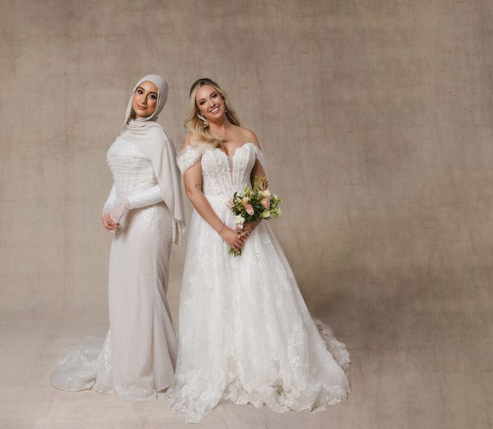 Two models pose against a neutral studio backdrop wearing wedding gowns. The model on the left wears Drew by Maggie Sottero, a fitted gown with shimmering embellishments and a coordinating hijab. The model on the right wears Skylar by Maggie Sottero, an off-the-shoulder lace ballgown featuring a structured bodice, full skirt, and floral bouquet.