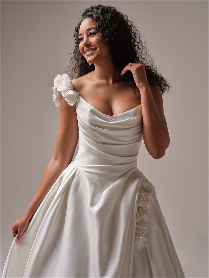 Bridal portrait of the Maggie Sottero Antoinette ballgown wedding dress in ivory, showcasing a draped strapless neckline, softly ruched satin bodice, dropped waist, full ballgown skirt, and dimensional floral accents at the shoulder and hip.