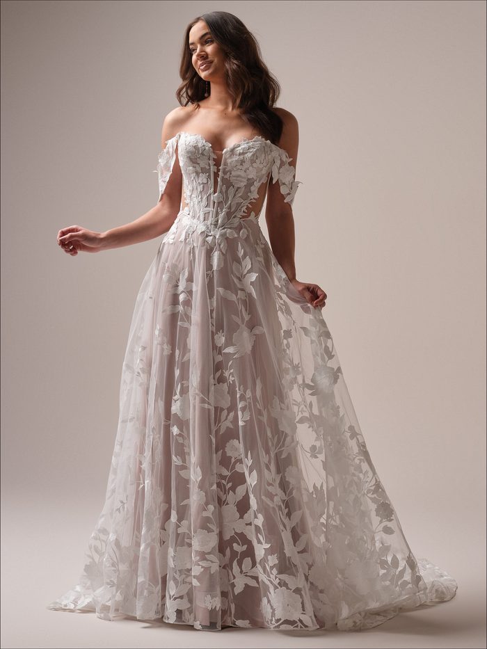 Natalie by Maggie Sottero A-line wedding dress featuring a plunging strapless neckline, detachable off-the-shoulder floral straps, and dimensional floral lace appliqués cascading over a soft blush-lined tulle skirt. Front view shown in a studio setting.