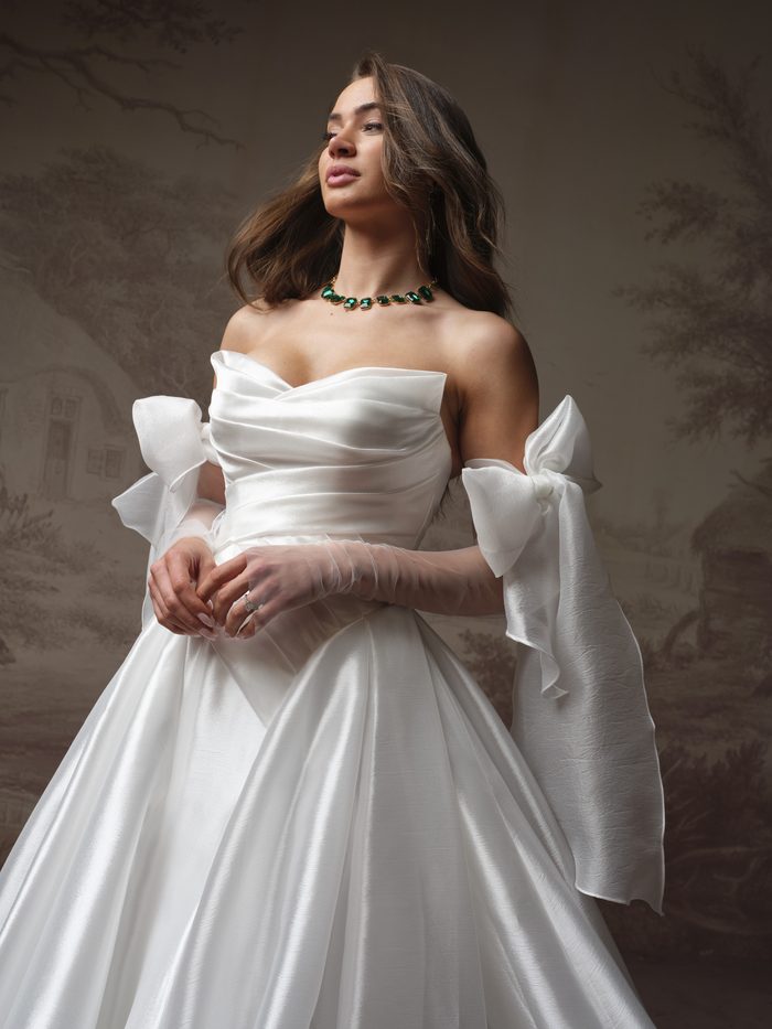 Bridal portrait of the Maggie Sottero Rosamund A-line wedding dress in ivory, featuring a draped strapless neckline, fitted satin bodice, full ballgown skirt, and detachable oversized bow sleeves worn off the shoulders.