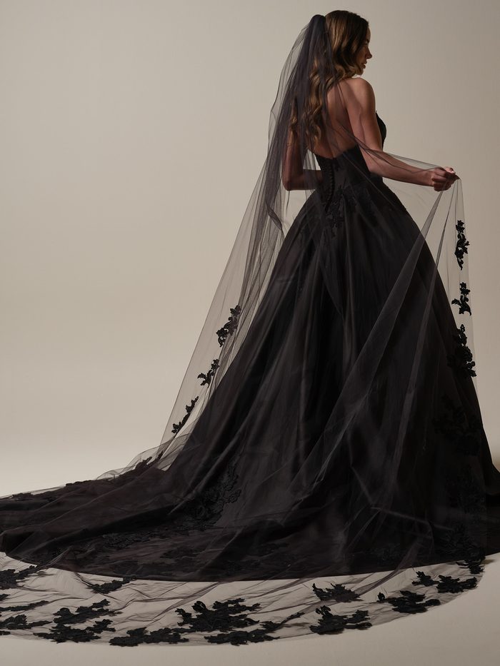 Back view of the Serafina by Maggie Sottero black ballgown wedding dress, highlighting the structured strapless bodice, voluminous skirt, and sweeping train. A cathedral-length black veil with floral lace appliqués cascades behind the gown, creating a dramatic silhouette in a studio setting.