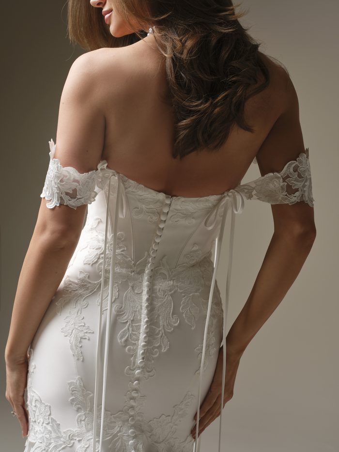 Back view of the Maggie Sottero Serafina Lane Fit-and-Flare wedding dress in ivory, showcasing off-the-shoulder lace sleeves, a button-back closure, delicate ribbon ties, and ornate floral lace appliqués accenting the fitted silhouette.