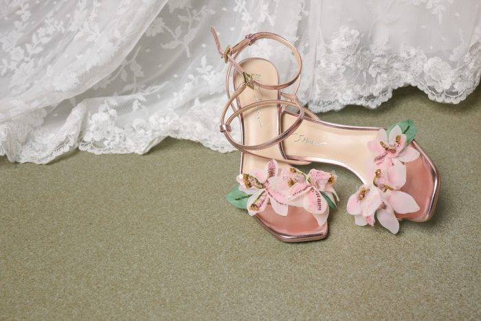 Pink metallic bridal heels featuring dimensional floral embellishments displayed beside the lace hem of a wedding dress.