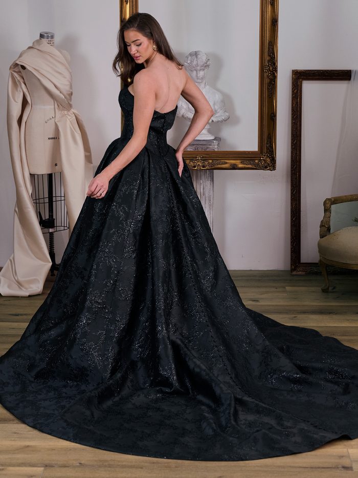 Back view of the Aberdeen by Sottero and Midgley ballgown wedding dress in black jacquard, showcasing a fitted strapless bodice, covered button detailing, and a sweeping cathedral-length train with subtle textured patterning. Displayed in a bridal studio with vintage décor.