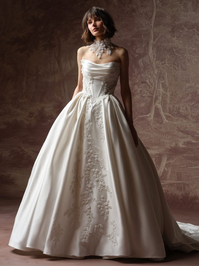 Burke Lane by Sottero and Midgley ballgown wedding dress featuring a draped strapless neckline, basque waist, and voluminous satin skirt with intricate floral appliqué accents. Front view showcases the dramatic silhouette and coordinating floral choker in a studio setting.