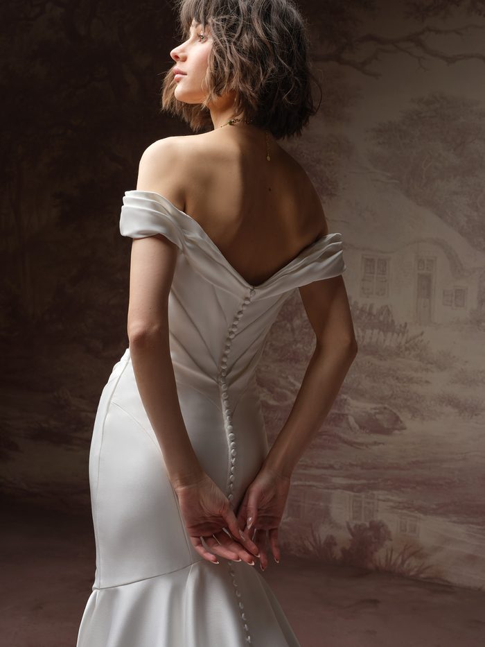 Back view of the Sottero and Midgley Recine fit-and-flare wedding dress in ivory, showcasing an off-the-shoulder neckline, low V-back, covered button closure, fitted crepe silhouette, and flared skirt with a chapel-length train.