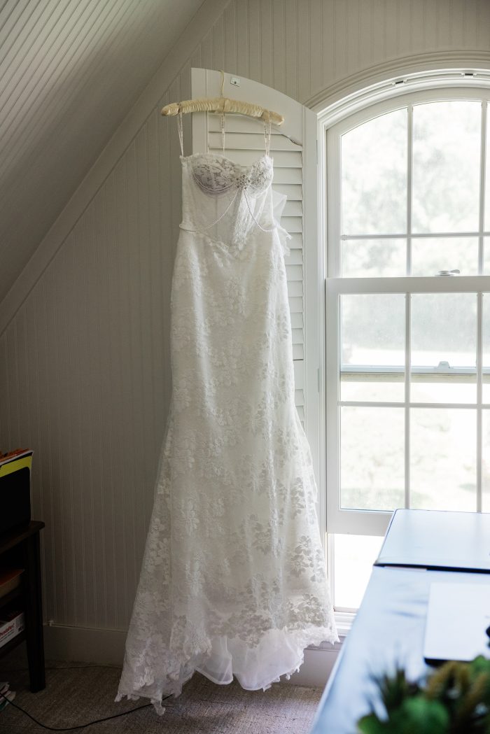 White floral lace wedding dress, Devon, hangs beside a sunlit window, featuring a sheer corset bodice, delicate spaghetti straps, draped pearl detailing, and a fitted silhouette with a flowing train.