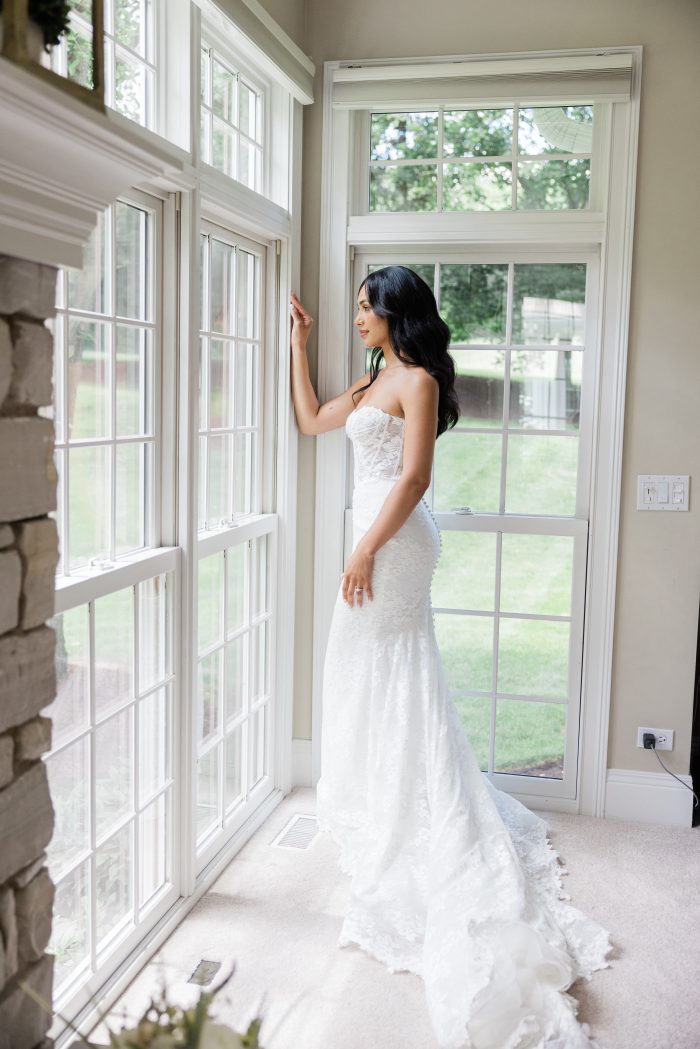 Bride poses beside a window in a strapless stretch lace wedding dress featuring a sheer corset bodice, figure-hugging silhouette, and flowing train.