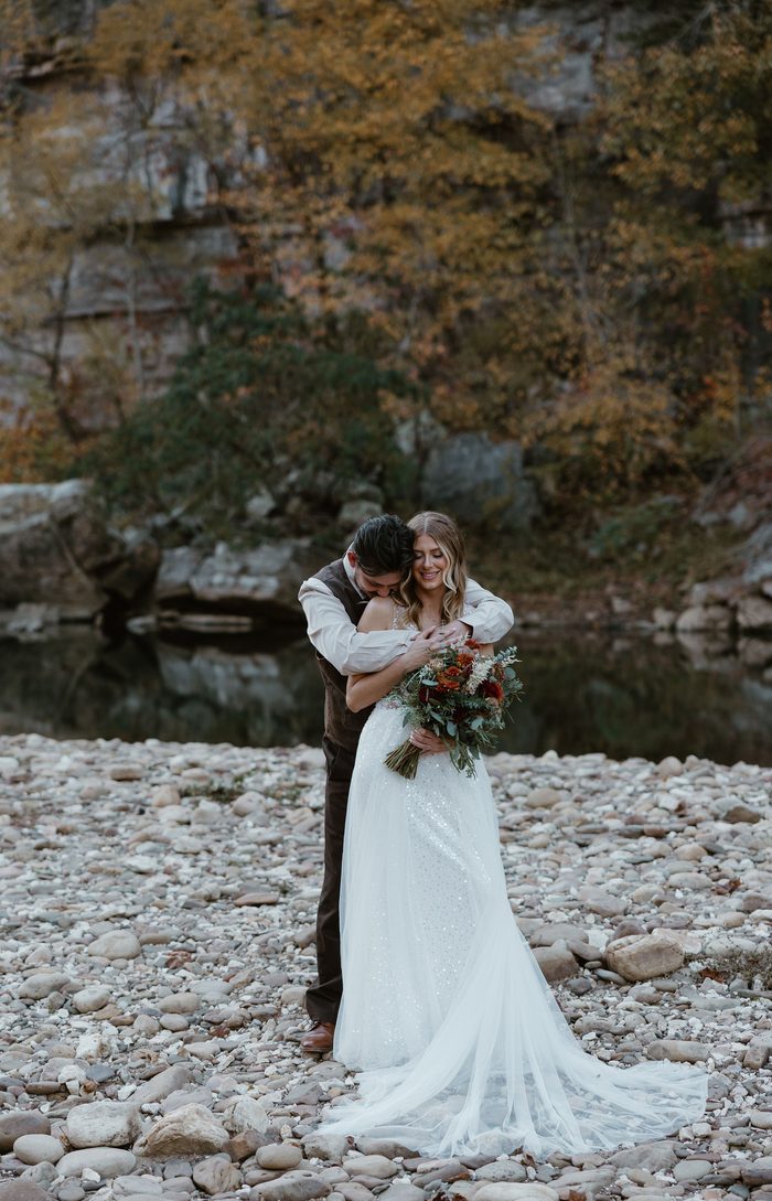 Bride and groom standing on a rocky riverside, with the bride wearing an ivory Maggie Sottero wedding dress featuring a fitted embellished bodice, shimmering glitter tulle A-line skirt, and flowing train. The couple poses beside calm water with autumn foliage and limestone cliffs in the background, while the bride holds a bouquet of rust, ivory, and greenery florals.