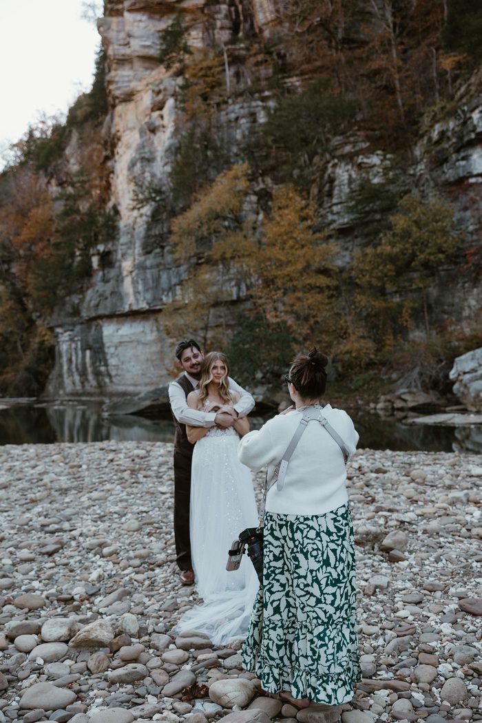 Behind-the-scenes photo of two photographers kneeling on a rocky riverside while photographing a couple in the distance. The bride wears an ivory Maggie Sottero wedding dress featuring a fitted embellished bodice, glitter tulle A-line skirt, and flowing train. Towering limestone cliffs, calm water, and autumn foliage create a scenic backdrop.