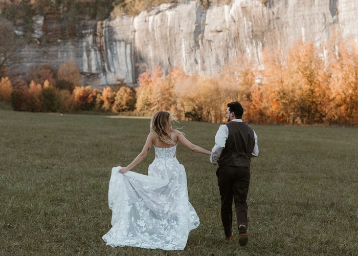 Rear view of a couple walking hand in hand across an open field beneath towering limestone cliffs and autumn foliage. The bride wears an ivory Natalie by Maggie Sottero wedding dress featuring a strapless floral lace bodice, sheer corset back, and flowing A-line skirt with oversized floral lace motifs. The bride lifts part of the skirt while walking, showcasing the gown's sweeping train and romantic movement.