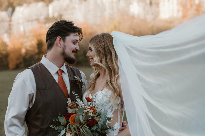 Close-up of a couple standing outdoors, with the bride wearing an ivory Natalie by Maggie Sottero wedding dress featuring a strapless floral lace bodice with sheer side panels. A flowing cathedral-length veil billows behind the bride while a bouquet of rust, burgundy, ivory, and greenery florals complements the warm autumn landscape in the background.