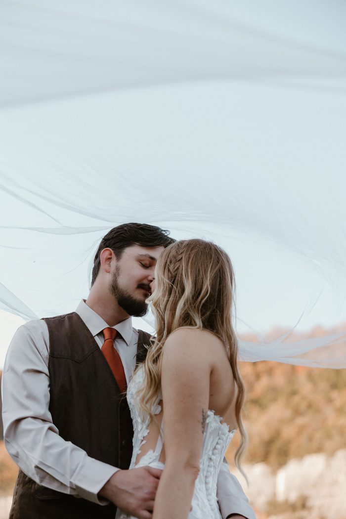 Close-up of a couple standing beneath a billowing cathedral-length veil outdoors. The bride wears Natalie by Maggie Sottero, an ivory A-line wedding dress with a strapless floral lace bodice, sheer side panels, and a structured corset back adorned with oversized botanical lace appliqués. Warm autumn colors and towering limestone cliffs frame the romantic portrait.