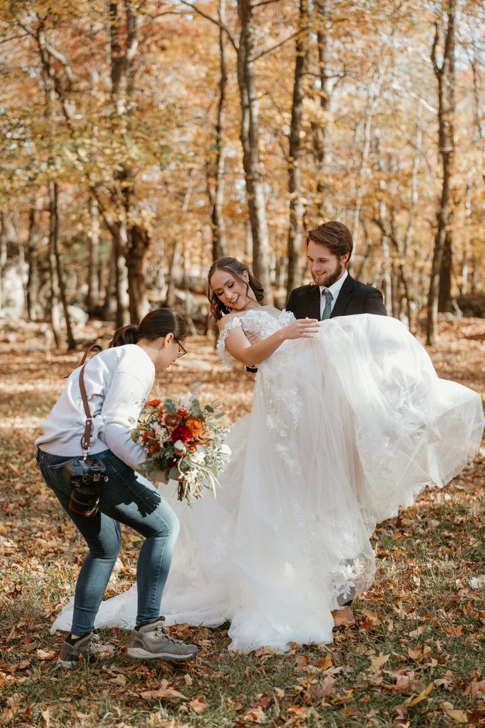 Behind-the-scenes photo of a photographer adjusting a bouquet while a couple poses in a sunlit forest. The bride wears Wicklow by Sottero and Midgley, an ivory A-line wedding dress with an off-the-shoulder floral lace bodice, textured lace appliqués, and a full layered tulle skirt. Autumn leaves blanket the ground beneath the woodland setting.