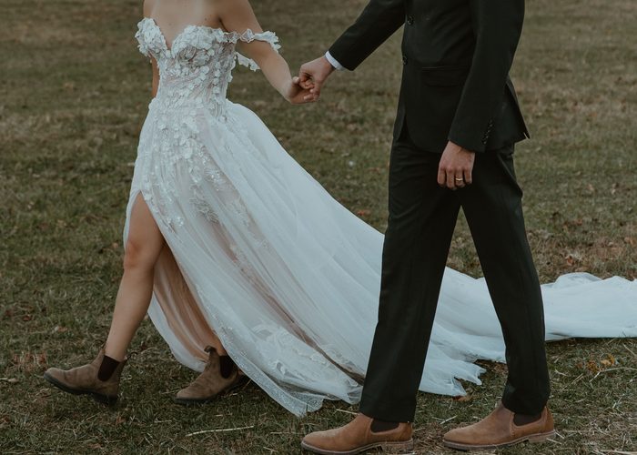 Close-up of a couple walking hand in hand across a grassy field. The bride wears Wicklow by Sottero and Midgley, an ivory A-line wedding dress featuring an off-the-shoulder floral lace bodice, dimensional lace appliqués, a sheer corset structure, and a thigh-high slit beneath a flowing glitter tulle overskirt. The sweeping train and layered skirt create dramatic movement as the couple walks together.
