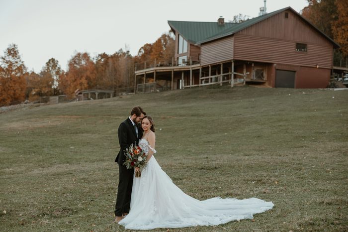 Couple posing on a grassy hillside with a rustic lodge and autumn trees in the background. The bride wears Wicklow by Sottero and Midgley, an ivory A-line wedding dress featuring an off-the-shoulder floral lace bodice, dimensional lace appliqués, and a flowing tulle skirt with a sweeping train. The bride holds a bouquet of rust, ivory, and greenery florals while standing beside a suited partner.