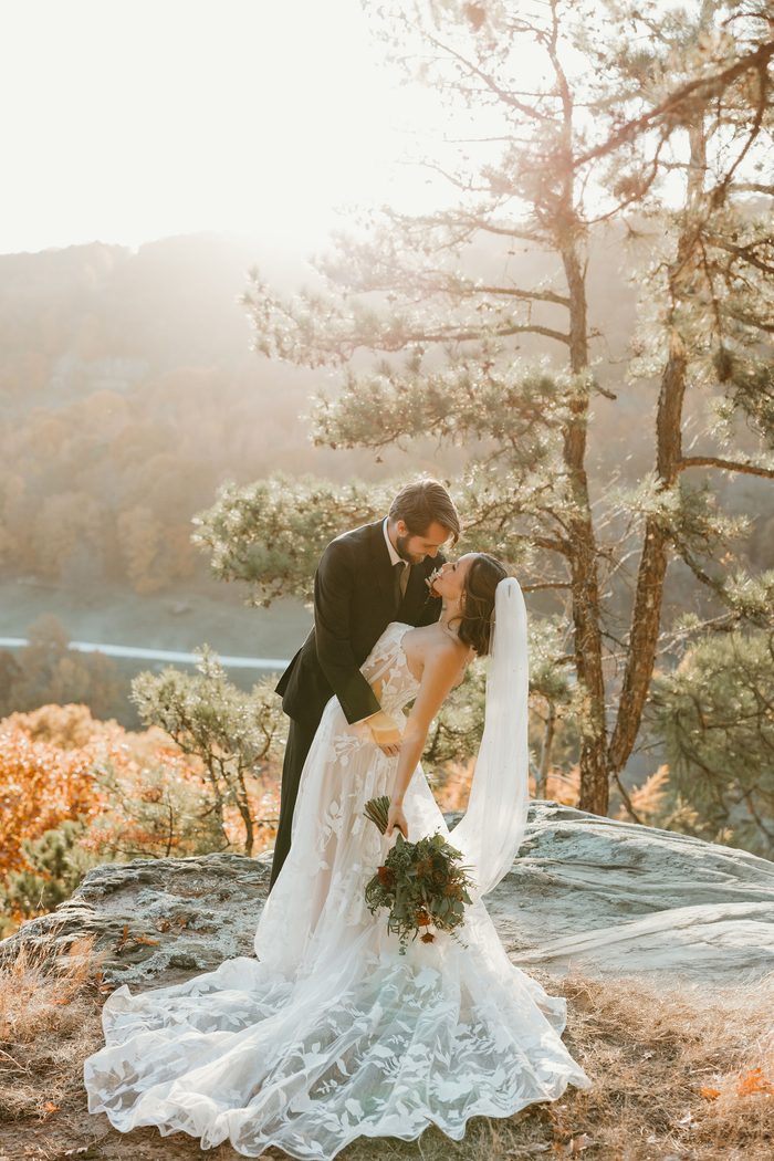 Couple posing on a rocky overlook surrounded by pine trees and autumn foliage. The bride wears Natalie by Maggie Sottero, an ivory A-line wedding dress featuring a strapless floral lace bodice, sheer corset detailing, and oversized botanical lace motifs throughout the skirt and train. A fingertip veil and a bouquet of rust, ivory, and greenery florals complete the look as the couple stands against a sunlit landscape backdrop.