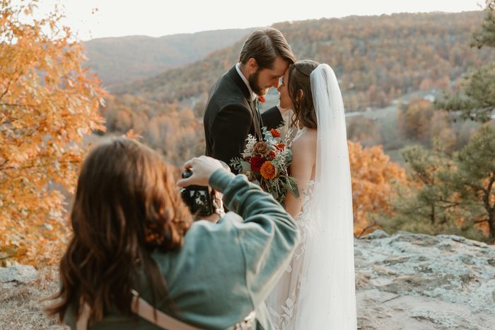 Behind-the-scenes photo of a photographer capturing a couple embracing on a rocky overlook surrounded by vibrant autumn foliage. The bride wears Natalie by Maggie Sottero, an ivory A-line wedding dress featuring a strapless floral lace bodice, sheer corset detailing, and oversized botanical lace motifs throughout the skirt and train. A pearl-detailed veil and a bouquet of rust, burgundy, ivory, and greenery florals complete the bridal look.