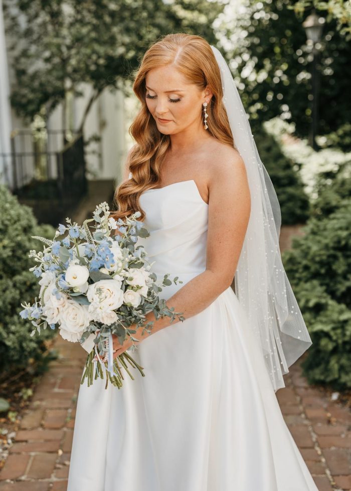 Bride in a strapless Mikado wedding dress with a draped neckline and flowing A-line skirt holds a bouquet of white and blue flowers, wearing a delicate pearl-dotted veil.