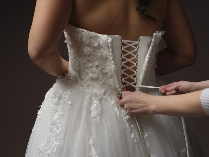 Close-up of a bride being laced into a floral lace wedding dress with a corset back and dimensional appliqués.