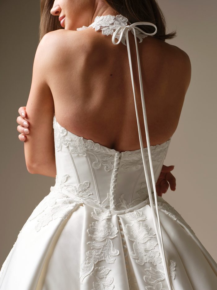 Back view of the Serafina Lane choker by Maggie Sottero, a band of embroidered floral lace tied at the nape with a thin satin ribbon whose long ends fall down the bride’s back to a covered-button ballgown with lace appliqués.