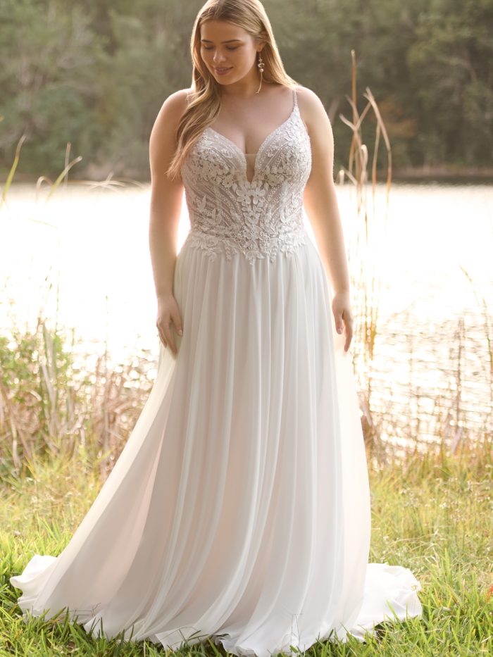 Bride poses by a sunlit lake in an organza A-line wedding dress featuring a plunging V-neckline, floral lace bodice, and flowing skirt.