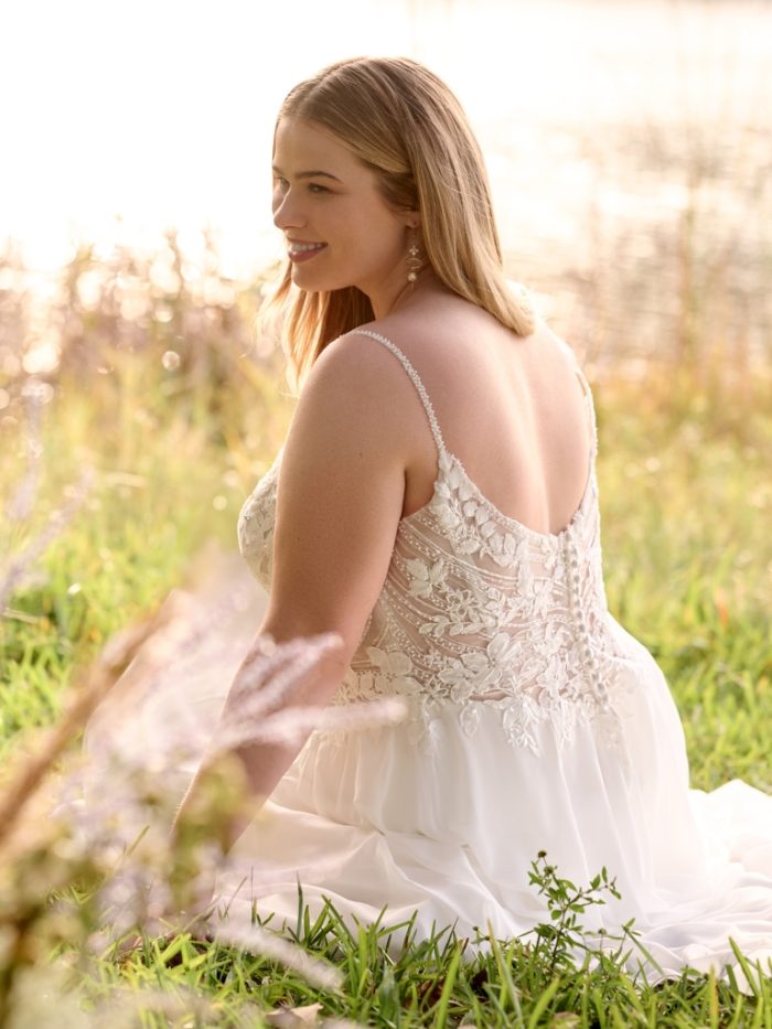 Bride sits in a grassy field wearing an organza A-line wedding dress with delicate floral lace appliqués, a sheer corset bodice, and thin beaded straps.