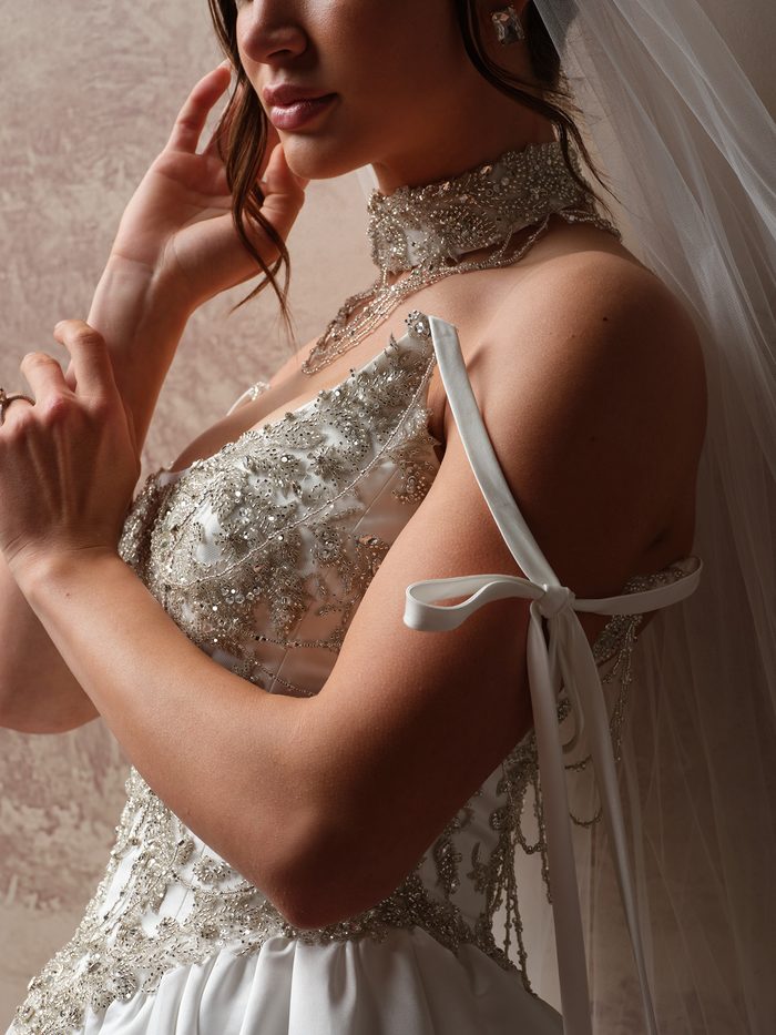 Side profile of a bride touching her face, showing the Aelarion wedding dress choker by Sottero and Midgley with its crystal-beaded band and draped strands, alongside the gown’s satin shoulder ribbon tied in a bow and its silver beaded bodice.