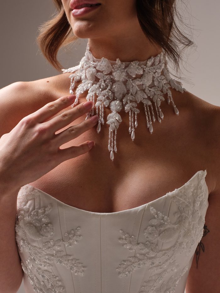 Close-up of the Amadora choker by Sottero and Midgley, with the bride’s hand resting below it. The high collar is covered in sequined and beaded floral leaves, with dangling strands of tiny beads ending in pearl teardrops above a beaded strapless corset.