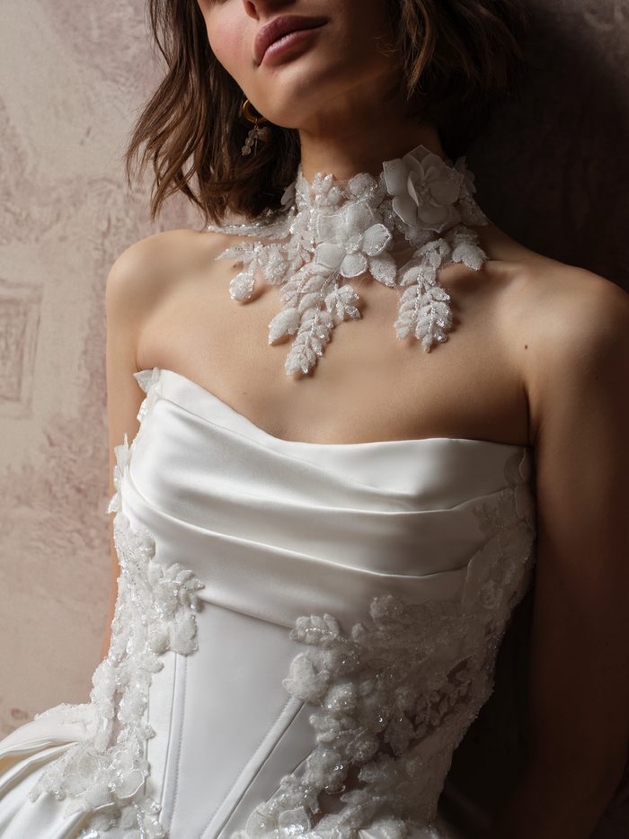 Close-up of the Burke bridal choker by Sottero and Midgley, showing its sheer high-neck base layered with dimensional white organza flowers and beaded, sequined leaves that fall in delicate sprays toward the neckline of a draped satin bodice.