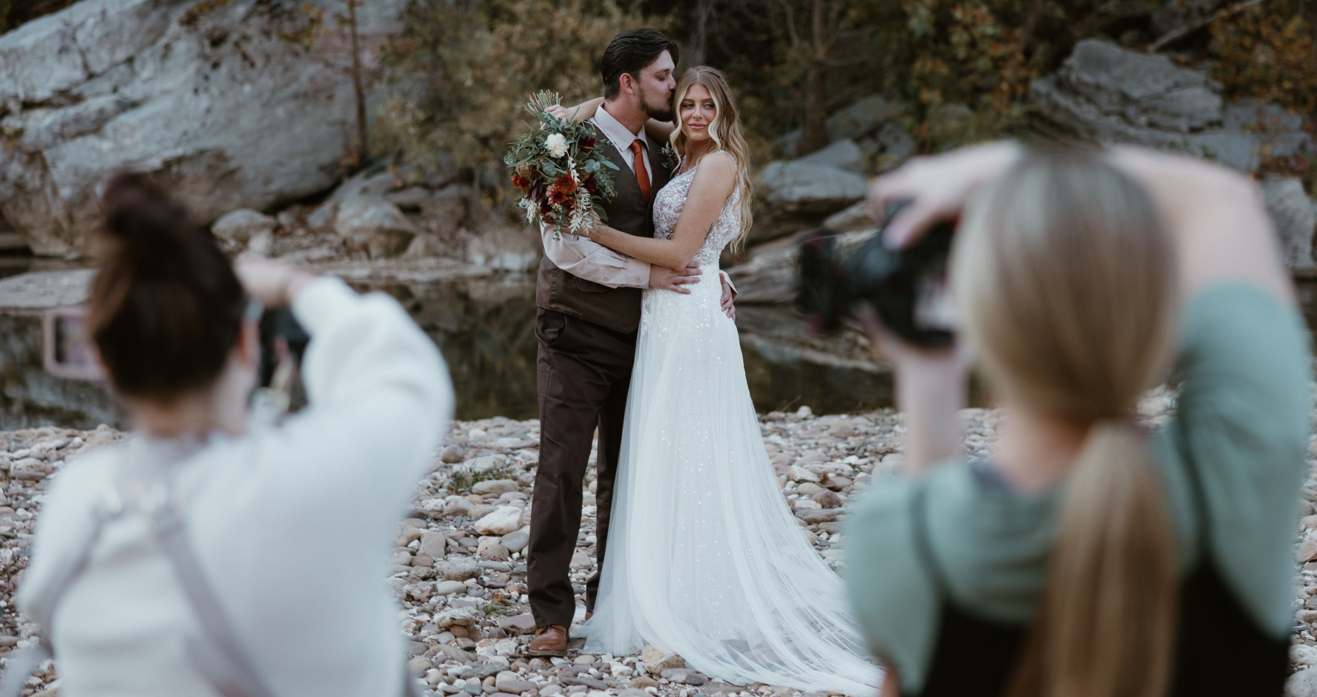 Behind-the-scenes photo of a bride and groom, the bride in a Maggie Sottero wedding dress posing beside a rocky riverside. The ivory A-line gown features a glitter tulle skirt, a fitted embellished bodice, and a flowing train. Two photographers frame the scene in the foreground while capturing the styled bridal portrait.